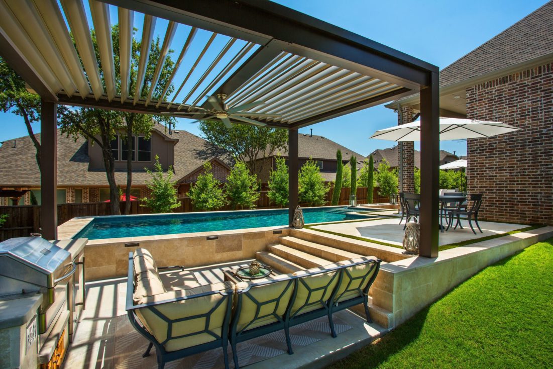 A motorized pergola with open louvers shades a residential seating area in a large backyard with green grass.
